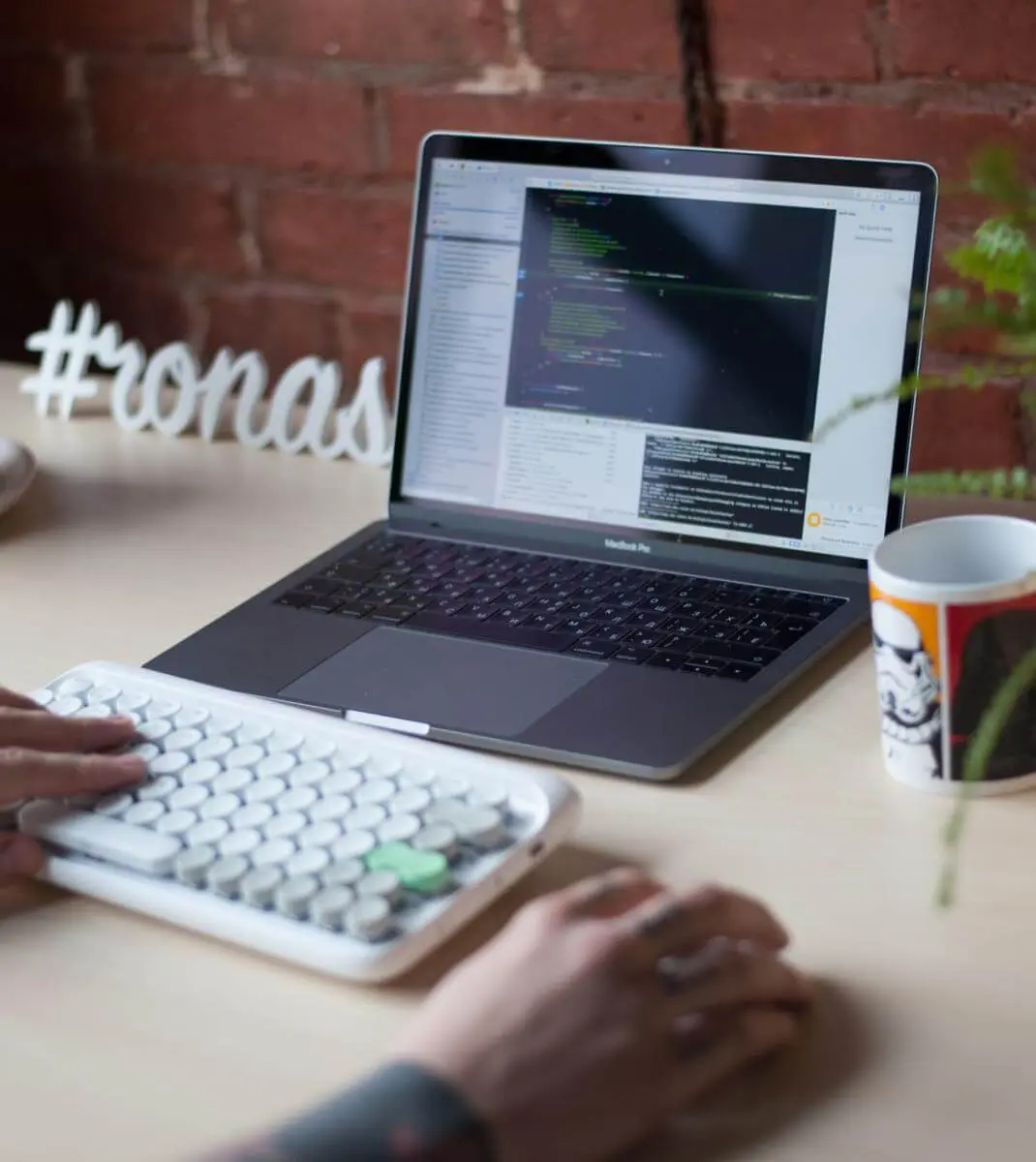 A developer’s hands typing on a keyboard in front of a laptop showing code, next to a Ronas IT sign and a mug