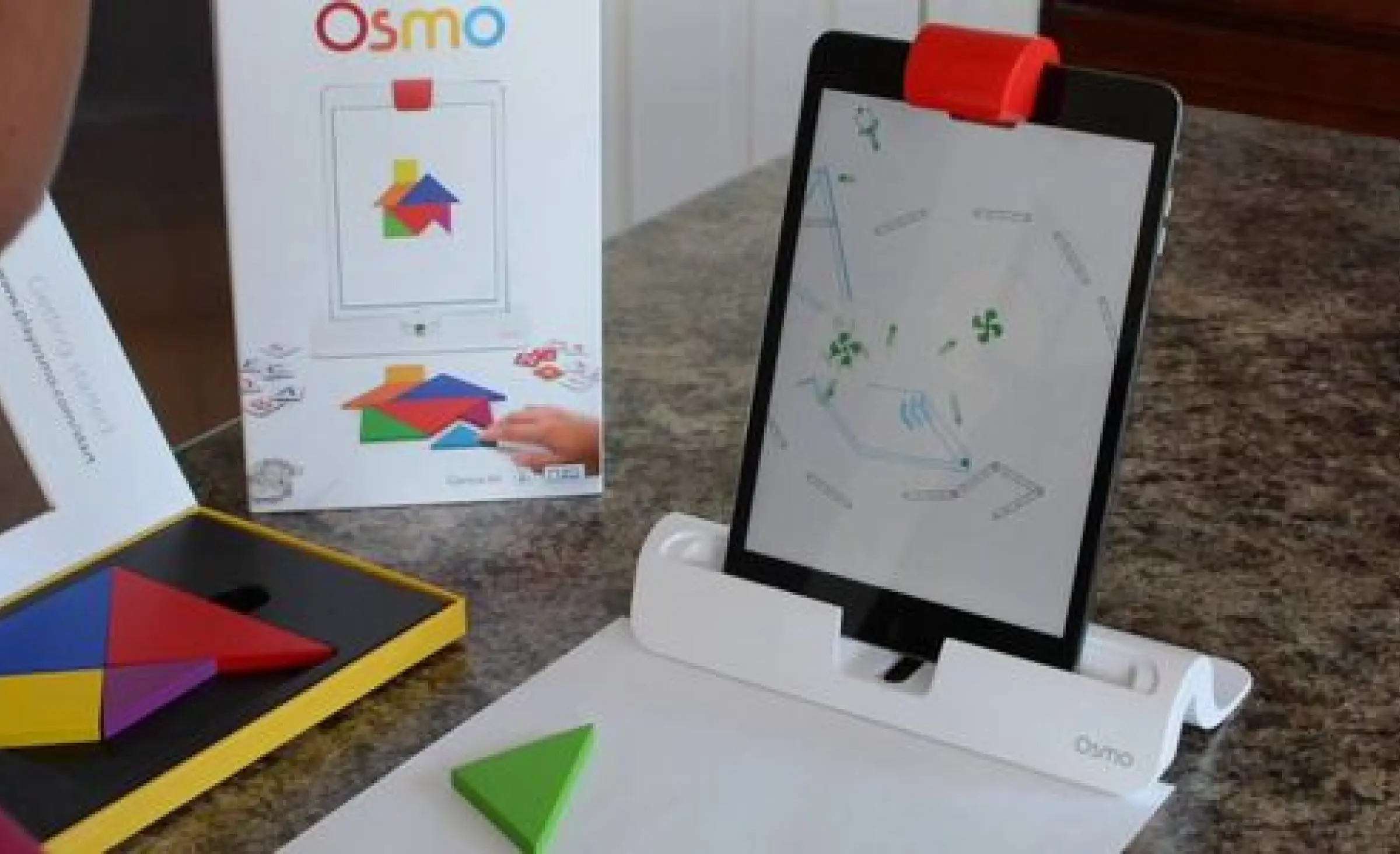 An Osmo kit on a table: a tablet in its base beside tangram pieces and a game box