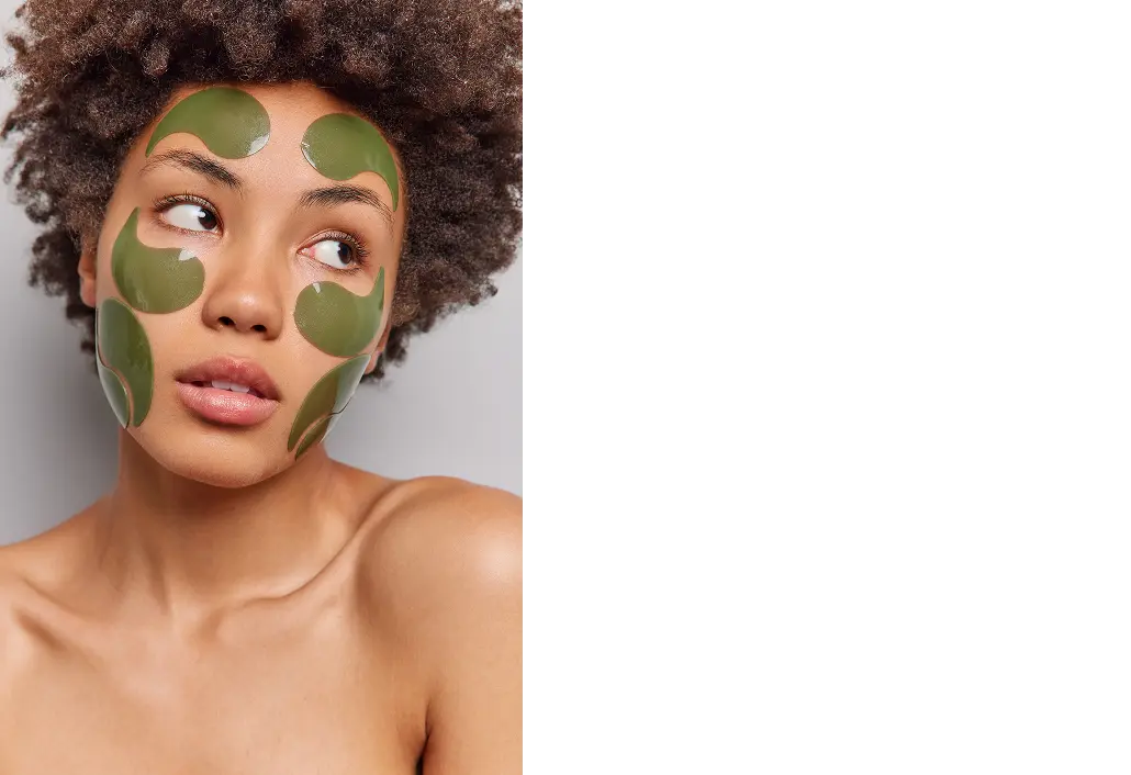 Woman with green clay face mask patches looking aside against a neutral background.