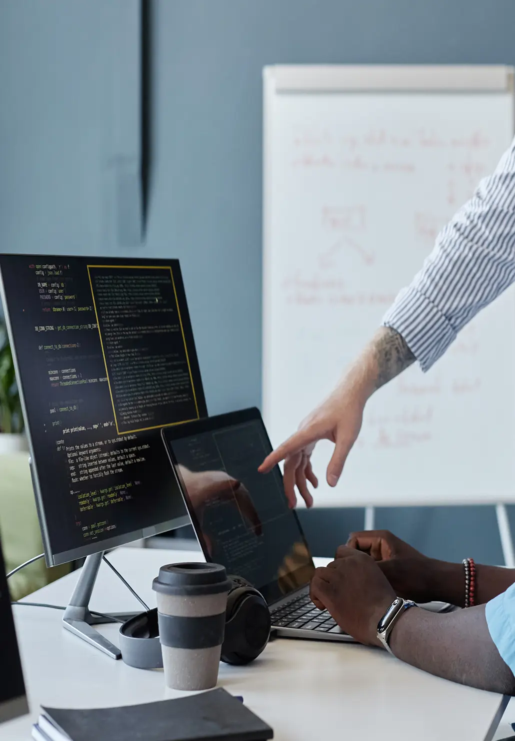 Developer pointing at code on a laptop screen while a colleague types, with a whiteboard in the background.