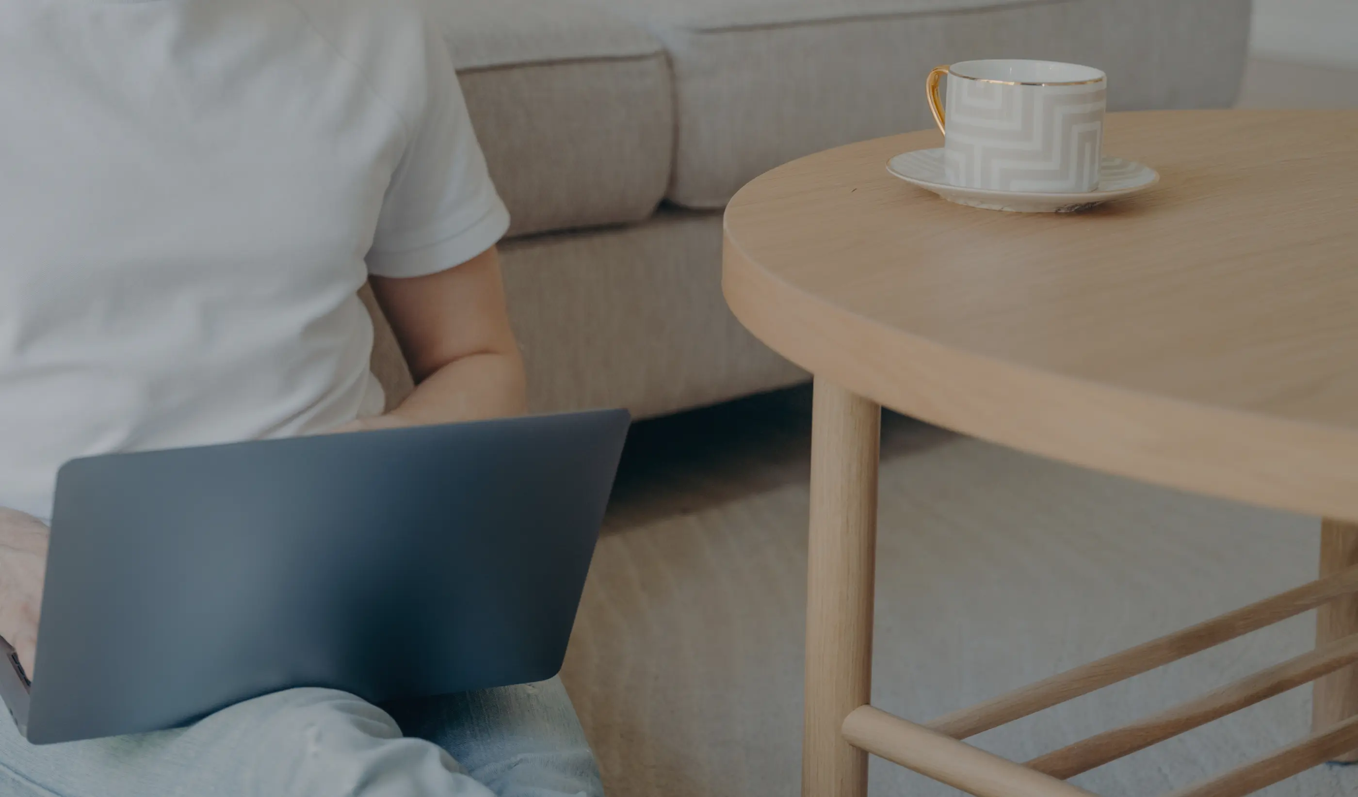 Person on a sofa using a laptop on their lap, next to a wooden table with a coffee cup.